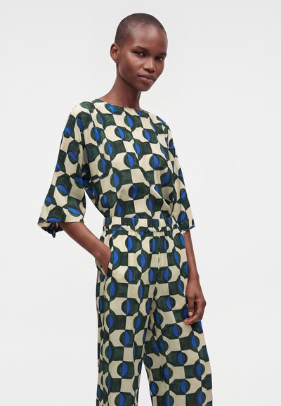 Femme portant un top ample et un pantalon assortis avec un motif géométrique vert et bleu, debout avec une main dans la poche.