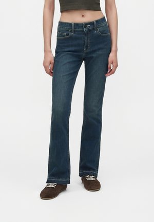 Person iført mørkeblå jeans med lige ben, brune snørebåndssko og en kort mørkegrøn top mod en hvid baggrund.