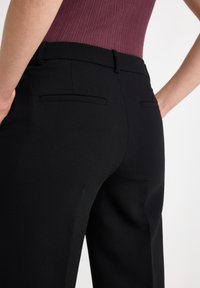 Schwarze maßgeschneiderte Hose mit glatter Textur, zwei Gesäßtaschen und einem mittelhohen Bund, kombiniert mit einem gerippten rotbraunen Oberteil.