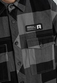 Schwarz-graues kariertes Hemd mit Knopfleiste, zwei Brusttaschen und einem rechteckigen REELL-Label auf der linken Seite.