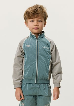 Niño pequeño con chaqueta con cremallera en azul verdoso y gris y pantalones a juego, de pie tranquilamente frente a un fondo liso.