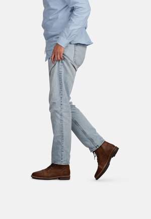 Braune Lederstiefel, leichtes blaues Hemd und hellblaue Jeans. Die Stiefel haben eine Schnürung; die Jeans sind schlank geschnitten mit einem klassischen Schnitt und leichtem Used-Look.