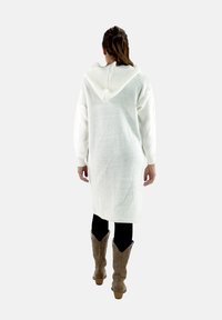 Sudadera larga blanca de punto con un bolsillo, textura acanalada y mangas largas, combinada con leggings negros y botas de vaquero marrones.