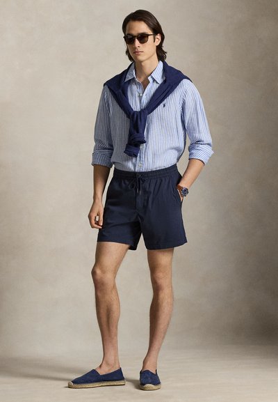 Σκούρα μπλε μαγιό σορτς με ελαστική μέση, συνδυασμένα με ανοιχτό γαλάζιο ριγέ πουκάμισο με κουμπιά και σκούρο μπλε πουλόβερ δεμένο γύρω από τους ώμους. Μπλε εσπαντρίγιες.