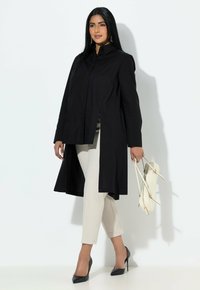 Czarna długa kurtka z wysokim kołnierzem, z bocznymi rozcięciami, w zestawie z kremowymi, zwężanymi spodniami i czarnymi szpiczastymi szpilkami, trzymająca jasną torbę.