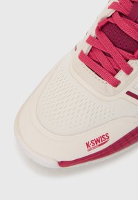 Lys beige idrettssko med røde detaljer, pustende mesh-overdel, teksturerte overflater og K-Swiss-merke på siden og sålen.