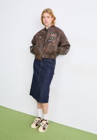 Brązowa skórzana kurtka bomberka z haftowanymi logo, noszona na ciemnowgranatowej spódnicy midi z denimu. Beżowo-brązowe sneakersy dopełniają strój. Zielona podłoga.