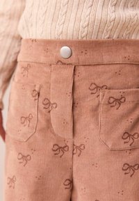 Pantaloni in corduroy di un morbido rosa, con una stampa di fiocchi discreta. Inclusi taschini frontali e una chiusura a scatto in metallo in vita.