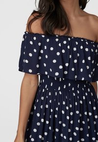 Žena v modré šaty s odhalenými rameny a bílými puntíky, se smyčkovým pasem, ukazující horní část trupu a paže.