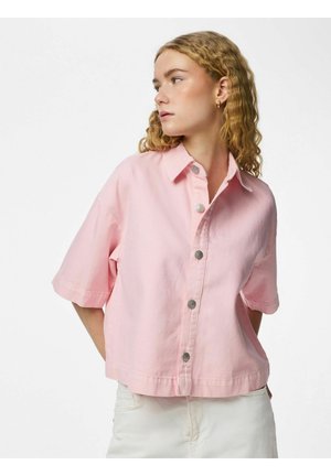 Mladá žena s kučeravými blond vlasmi, oblečená v voľnej ružovej krátkorukávovej košeli na gombíky a bielych nohaviciach, pozerajúca sa bokom.