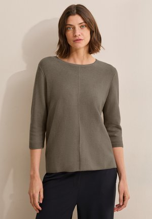 Graues Strickoberteil mit rundem Ausschnitt und drei Viertelärmeln. Hat einen geraden Schnitt und eine dezente Textur. Getragen mit dunklen Hosen.