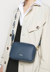 Blå läder crossbody-väska med texturerad yta, gyllene detaljer och ett mönstrat band med en logotyp. Bärs över en beige kappa.