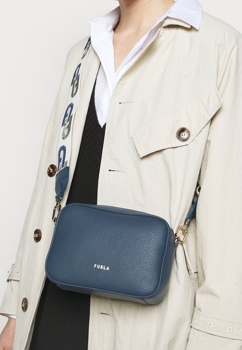 Blå läder crossbody-väska med texturerad yta, gyllene detaljer och ett mönstrat band med en logotyp. Bärs över en beige kappa.