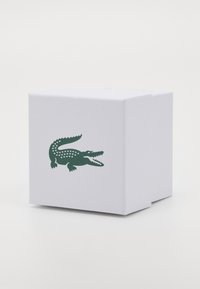 Valge ruudukujuline kast, mis on valmistatud tekstuuri materialist, mille ühel küljel on tume roheline krokodilli logo. Kast on mõeldud toote pakendamiseks.