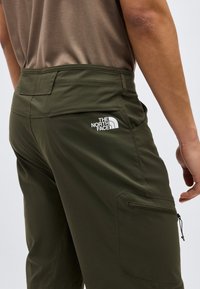 Άνδρας που φοράει χακί παντελόνι North Face με φερμουάρ στην πλαϊνή τσέπη και μπεζ πουκάμισο, εμφανιζόμενος από τη μέση πίσω έως το μέσο του μηρού.