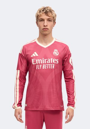 Ung mand med kort blondt hår iført en langærmet rød Real Madrid fodboldtrøje med hvide logoer og striber, vendt mod fronten.