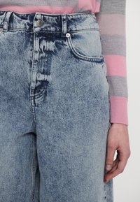 Light blue denim jeans with a faded texture, high-waisted design, front pockets, and metal button closure; paired with a striped sweater.
