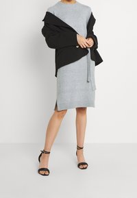 Abito grigio in maglia con scollatura rotonda e spacchi laterali, abbinato a un giubbotto nero oversize. Sandali neri con cinturini completano il look.