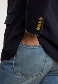 Blazer azul-marinho com quatro botões dourados na manga, apresentando um forro interior às riscas. Combinado com jeans de ganga azul clara que mostram o bolso traseiro.