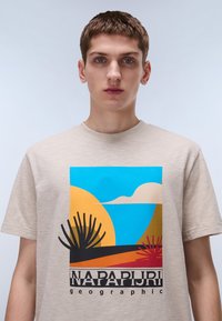 Bež majica s pisanom grafiko, ki vključuje elemente puščave, vključno z modrim nebom, soncem, gorami in abstraktno vegetacijo.