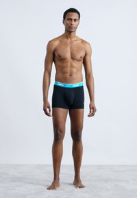 Boxers noirs avec une taille en logo turquoise de Nike, en tissu lisse, à coupe ajustée et longueur mi-cuisse. Le modèle est debout pieds nus.