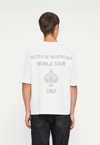 Witte katoenen t-shirt met grote grijze opdruk op de achterkant: "NOTES OF NOSTALGIA WORLD TOUR," een decoratief klavertje en "1962."