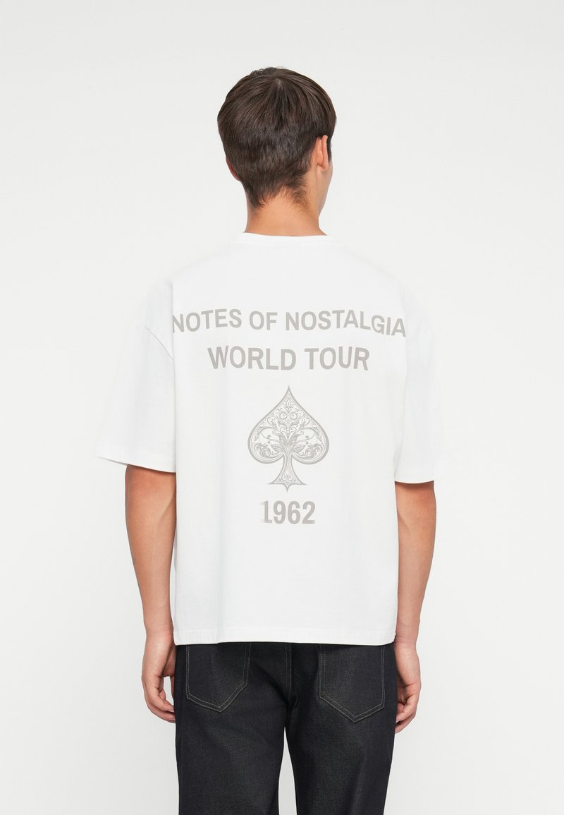 Witte katoenen t-shirt met grote grijze opdruk op de achterkant: "NOTES OF NOSTALGIA WORLD TOUR," een decoratief klavertje en "1962."