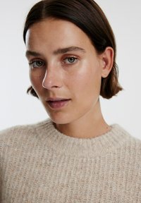 Lichtbeige gebreid sweater met een geribbelde textuur en een ronde halslijn, gedragen door een persoon met kort bruin haar en naturelle make-up.