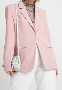 Light pink blazer with structured design, two front pockets, and single button closure, paired with a silver mini bag and patterned top.