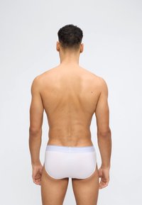 Άνδρας μοντέλο εμφανίζεται από πίσω φορώντας απλά λευκά εσώρουχα, στέκεται μπροστά σε απλό ανοιχτό γκρι φόντο.