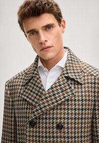 Brauner und beiger Hahnentritt-Mantel mit zweireihigem Design, großen Revers und dunklen Knöpfen, getragen über einem weißen Hemd.