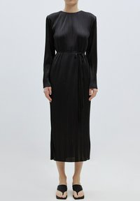 Schwarzes mittellanges Kleid mit langen Ärmeln, plissierter Textur, Rundhalsausschnitt und einem gebundenen Taillengürtel, kombiniert mit flachen schwarzen Sandalen.