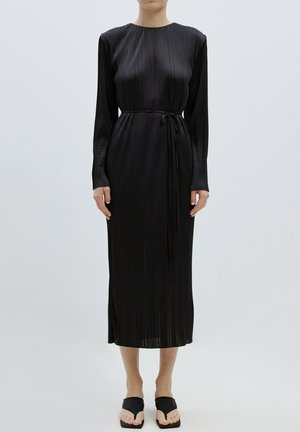 Schwarzes mittellanges Kleid mit langen Ärmeln, plissierter Textur, Rundhalsausschnitt und einem gebundenen Taillengürtel, kombiniert mit flachen schwarzen Sandalen.