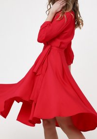 Robe rouge fluide en tissu léger, avec une taille cintrée par une ceinture, des manches longues et un ourlet évasé. Texture lisse et couleur vibrante.