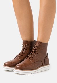 Bottines en cuir marron avec des détails de brogue, bout rond et semelle en caoutchouc blanc. Présente un design à lacets et une surface texturée.