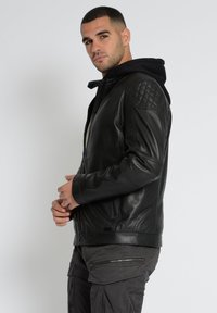 Schwarze Lederjacke mit gesteppten Schulterdetails und Kapuze. Verfügt über einen vorderen Reißverschluss und Seitentaschen, vor einem weißen Hintergrund.