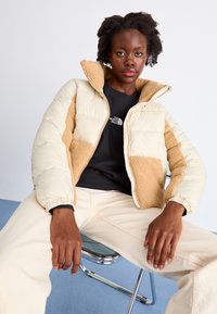 Opblaasjack met crèmekleurige en tankleurige panelen, gewatteerd patroon, fleece accenten aan de voorkant en een ritssluiting. Zwarte shirt eronder.