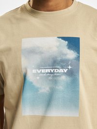 Beige bavlněné tričko s krátkým rukávem a kulatým výstřihem, na kterém je obdélníková modro-bílá grafika s mraky a textem "EVERYDAY."