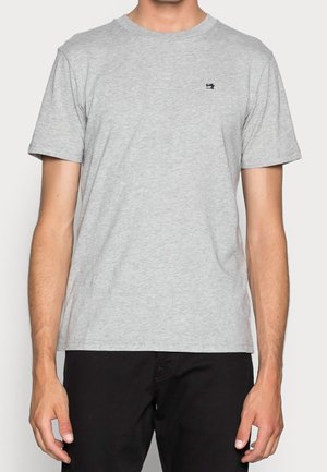 Mężczyzna ubrany w jasnoszary T-shirt z krótkim rękawem z małym ciemnym logo maszyny do szycia na lewej piersi oraz czarne spodnie.