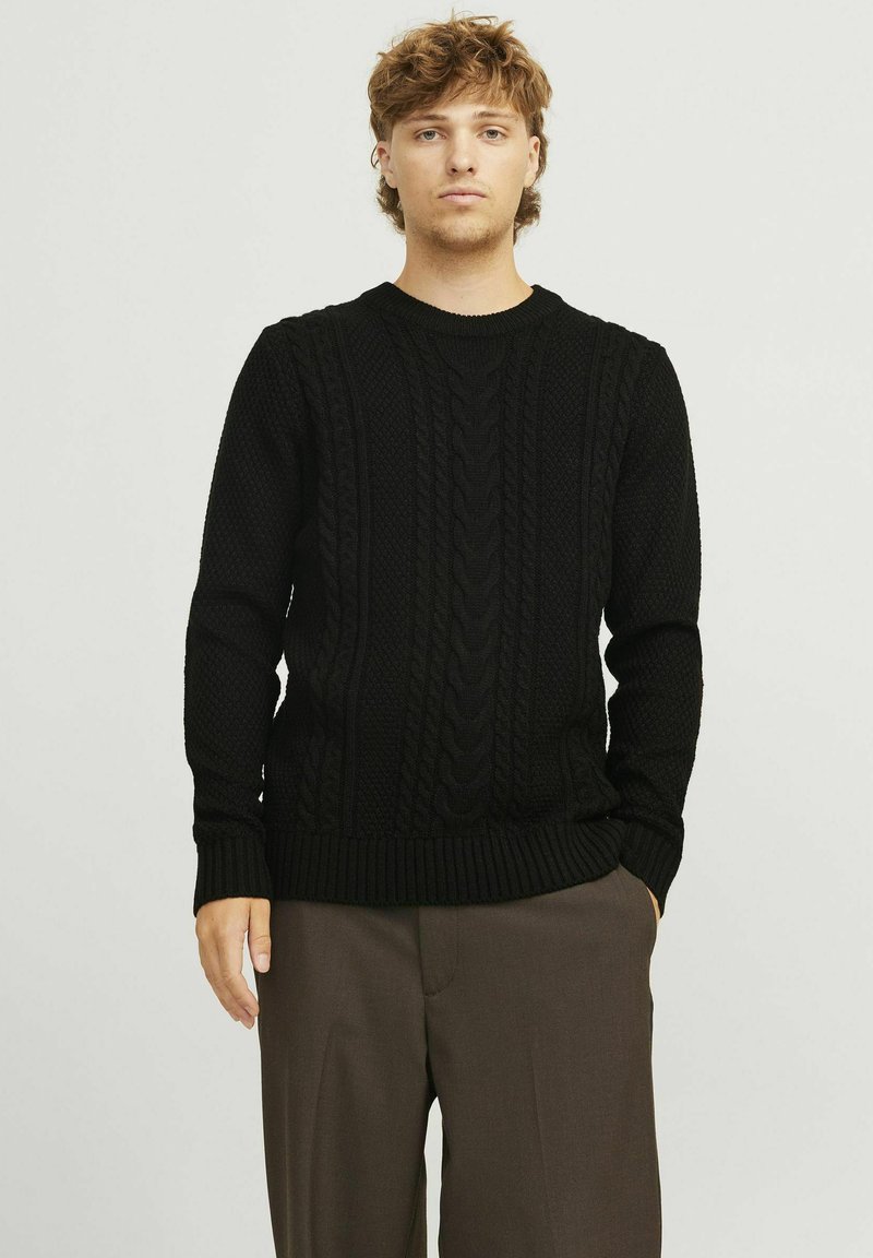 Schwarzer gestrickter Pullover mit Kabelmuster, gerippten Bündchen und Saum sowie Rundhalsausschnitt. Getragen über braunen Hosen vor einem schlichten Hintergrund.