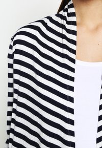 Persona que lleva un cárdigan de rayas horizontales en blanco y negro sobre una camiseta blanca, mostrado desde el hombro hasta el pecho.