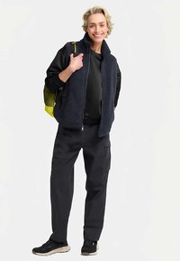 Navy fleece vest over a black top, paired with dark cargo pants and black sneakers, holding a yellow mesh backpack. Casual design and fit.