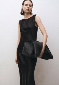 Schwarzes ärmelloses Kleid aus glattem, seidenähnlichem Stoff mit taillierter Silhouette; hält eine weiche schwarze Lederhandtasche mit gestepptem Muster.