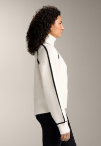 Maglione a collo alto bianco e soffice con strisce verticali nere lungo le maniche. Tessuto morbido, vestibilità rilassata e polsini a coste.