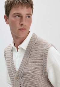 Hellbeige gestrickte Weste mit V-Ausschnitt, die ein strukturiertes Fischgrätmuster und einen Knopf auf der Vorderseite aufweist. Trägt man über einem weißen Hemd mit Kragen.