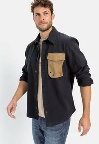 Marine shirtjack met een beige borstzak, lange mouwen opgerold, knoopsluiting en een textuur van stof; gedragen over een beige shirt.