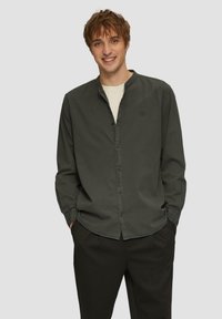 Camicia button-up verde scuro con colletto alla mandarino, maniche lunghe e un logo discreto sul petto, abbinata a pantaloni neri.