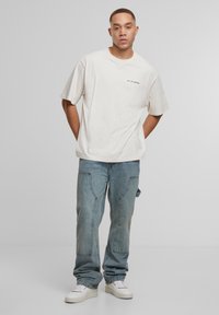 Übergroßes cremefarbiges T-Shirt aus Baumwolle mit kurzen Ärmeln und dem Schriftzug "2Y STUDIOS". Kombiniert mit hellblauen, weiten Jeans und weißen Turnschuhen.