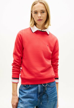 Giovane che indossa una felpa rossa con collo a giro sopra una camicia bianca con colletto e jeans blu con una cintura a corda nera infilata nei passanti.