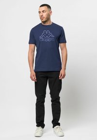Sötétkék Kappa póló nagy, sötétkék logóval a mellkason. Fekete farmernadrággal és fehér tornacipővel párosítva, kerek nyakú dizájnnal.